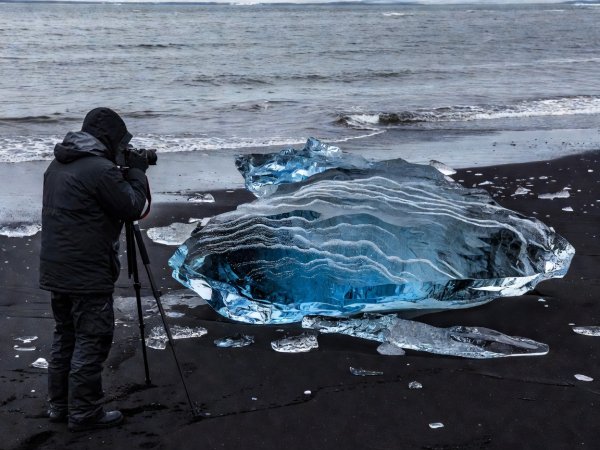 Photographer framing a stranded dense blue glacial ice block stranded on dark volcanic basalt sand